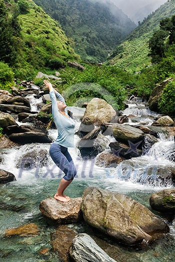 Young sporty fit woman doing yoga asana Utkatasana (chair pose) outdoors at tropical waterfall standing on stone Young sporty fit woman doing yoga asana Utkatasana (chair pose) outdoors at tropical waterfall standing on stone
