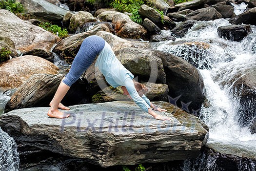 Young sporty fit woman doing yoga asana Adho mukha svanasana - downward facing dog - at tropical waterfall