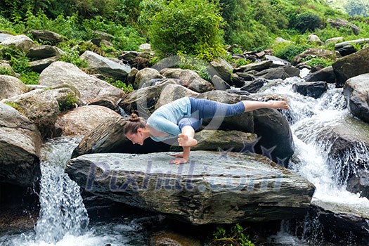 Young sporty fit woman doing yoga asana Eka Pada Koundinyasana 1 at tropical waterfall