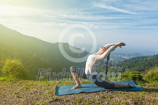 Sporty fit woman practices yoga Anjaneyasana - low crescent lunge pose outdoors in mountains in morning