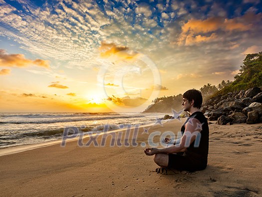 Young sporty fit man doing yoga meditating in padmasana lotus pose on tropical beach on sunset