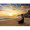 Young sporty fit man doing yoga meditating in padmasana lotus pose on tropical beach on sunset