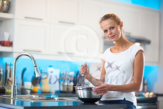 Pretty, young woman in her modern, clean and bright kitchen, fixing lunch/cooking/baking/making pancakes (color toned imagey; shallow DOF)