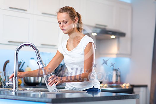 Pretty, young woman in her modern, clean and bright kitchen, pouring herself and drinking a glass of cold tap water(color toned imagey; shallow DOF)