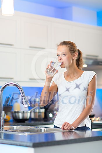 Pretty, young woman in her modern, clean and bright kitchen, pouring herself and drinking a glass of cold tap water(color toned imagey; shallow DOF)