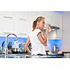 Pretty, young woman in her modern, clean and bright kitchen, pouring herself and drinking a glass of cold tap water(color toned imagey; shallow DOF)