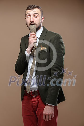 Young stylish man in a green suit and orange jeans with a tie in the mouth. Beige background.