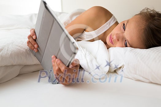 Pretty, young woman with a tablet computer in her bed, reading news, answering to emails, checking the weather forecast so that she nows what to wear today