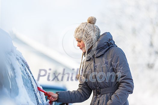 Young woman cleaning her car from snow and frost on a winter morning, she is freezing and needs to get to work