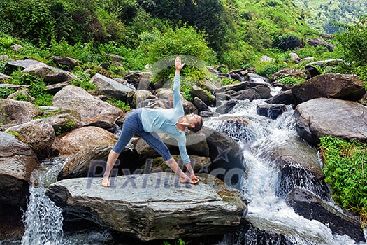Woman doing Ashtanga Vinyasa yoga asana Utthita trikonasana - extended triangle pose outdoors at waterfall in Himalayas