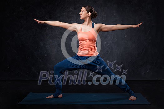 Beautiful sporty fit yogini woman practices yoga asana Virabhadrasana 2 - warrior pose 2 on dark background