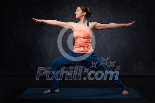 Beautiful sporty fit yogini woman practices yoga asana Virabhadrasana 2 - warrior pose 2 on dark background Beautiful sporty fit yogini woman practices yoga asana Virabhadrasana 2 - warrior pose 2 on dark background