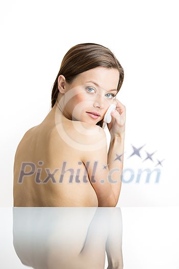 Skin care concept - Pretty, young woman removing face makeup with cotton swab pad - beautiful Caucasian model with perfect skin. Girl isolated on white background with some reflection underneath her (shallow DOF; color toned image)