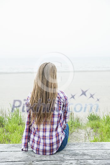 Back view of young woman with long blond hair sitting on old beach picnic table facing ocean wearing plaid shirt Back view of young woman with long blond hair sitting on old beach picnic table facing ocean wearing plaid shirt