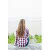 Back view of young woman with long blond hair sitting on old beach picnic table facing ocean wearing plaid shirt