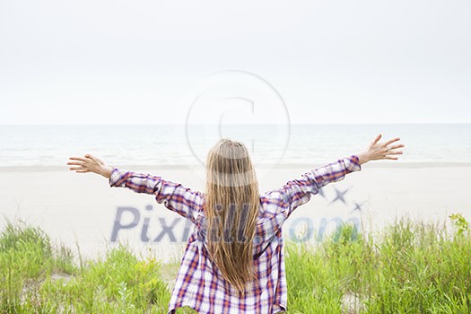 Back view of young woman with long blond hair and outstretched arms on empty beach facing ocean wearing plaid shirt Back view of young woman with long blond hair and outstretched arms on empty beach facing ocean wearing plaid shirt
