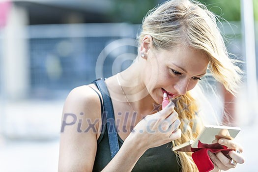 Beautiful young blonde woman applying lipstick with compact makeup mirror outside on city street