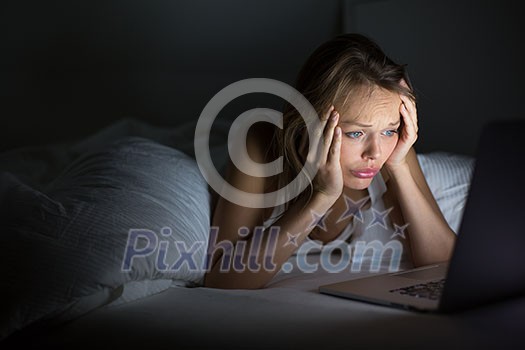 Pretty young woman watching something awful/sad on her laptop in bed at night in a dark bedroom (shallow DOF; color toned image)