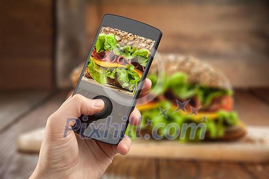 Hand taking photo of sandwich with smatphone on the wooden table with slices of tomatoes, ham, cheese and lettuce