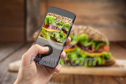 Hand taking photo of sandwich with smatphone on the wooden table with slices of tomatoes, ham, cheese and lettuce Hand taking photo of sandwich with smatphone on the wooden table with slices of tomatoes, ham, cheese and lettuce