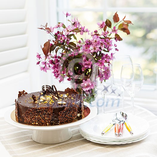 Round gourmet chocolate cake on table next to window with plates, cutlery, and pink spring bouquet
