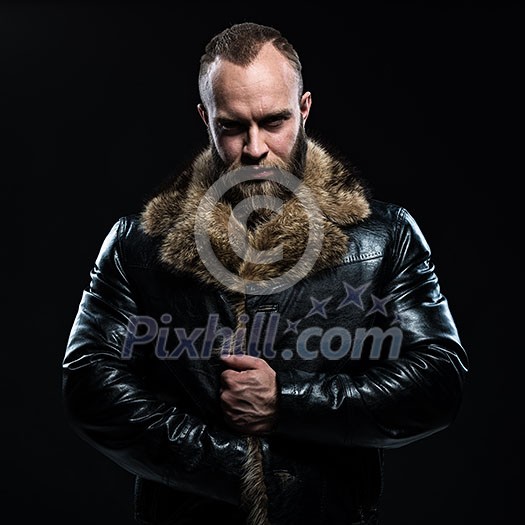 Brutal handsome glum unshaven man with beard and moustache in black fur coat with collar over dark background.