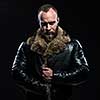 Brutal handsome glum unshaven man with beard and moustache in black fur coat with collar over dark background.