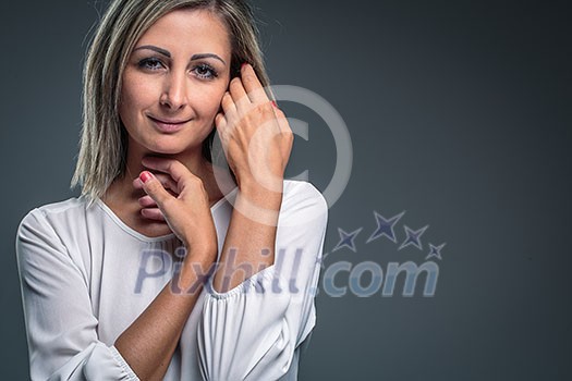 Portrait of a very attractive blonde, young woman on grey background, touching her lovely hair, looking both relaxed and confident (color toned image)