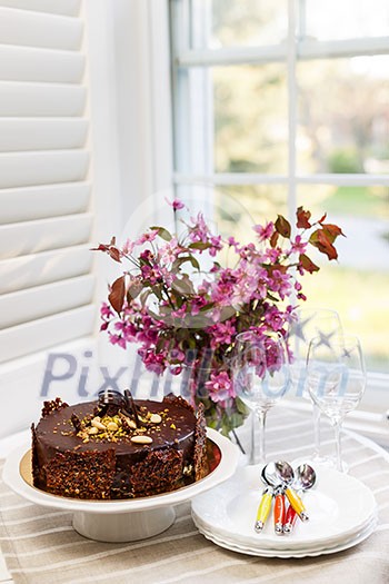 Round gourmet chocolate cake on table next to window with plates, cutlery, and pink spring bouquet