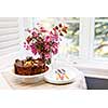 Round gourmet chocolate cake on table next to window with plates, cutlery, and pink spring bouquet