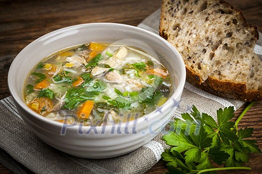 Chicken rice soup with vegetables in bowl and bread on rustic table