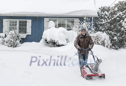 Man using snowblower to clear deep snow on driveway near residential house after heavy snowfall.