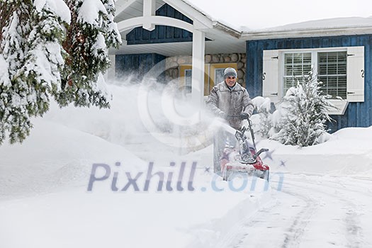 Man using snowblower to clear deep snow on driveway near residential house after heavy snowfall.