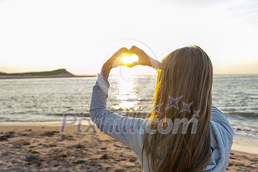 Blonde young girl holding hands in heart shape framing setting sun at sunset on ocean beach