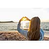 Blonde young girl holding hands in heart shape framing setting sun at sunset on ocean beach