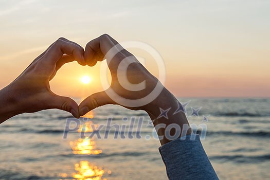 Hands and fingers in heart shape framing setting sun at sunset over ocean
