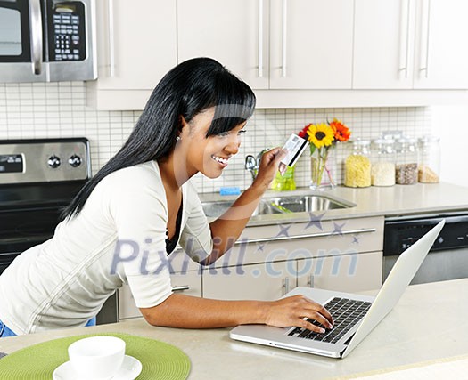 Smiling black woman online shopping using computer and credit card in kitchen Smiling black woman online shopping using computer and credit card in kitchen