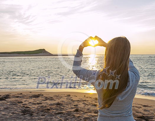 Blonde young girl holding hands in heart shape framing setting sun at sunset on ocean beach