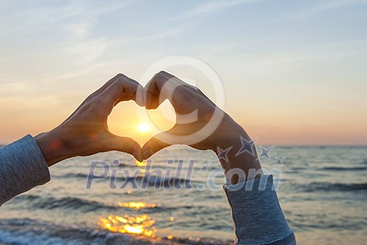 Hands and fingers in heart shape framing setting sun at sunset over ocean Hands and fingers in heart shape framing setting sun at sunset over ocean