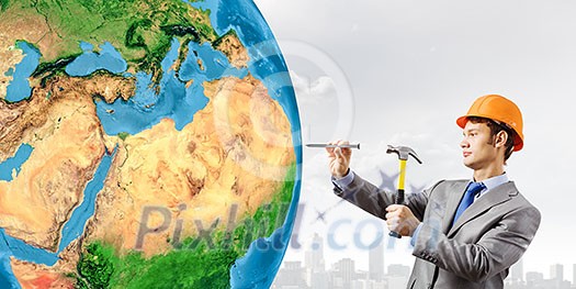 Businessman in helmet hammering nail in Earth planet. Elements of this image are furnished by NASA