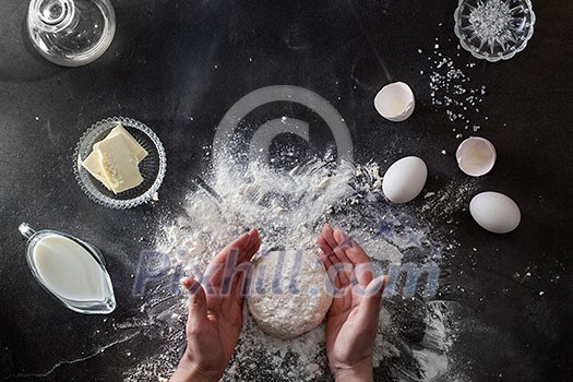 Woman's hands knead dough on table with flour and ingridients. Top view.