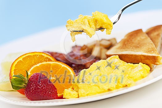 Scrambled eggs on a fork above breakfast plate with fruits toast and bacon