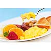 Scrambled eggs on a fork above breakfast plate with fruits toast and bacon