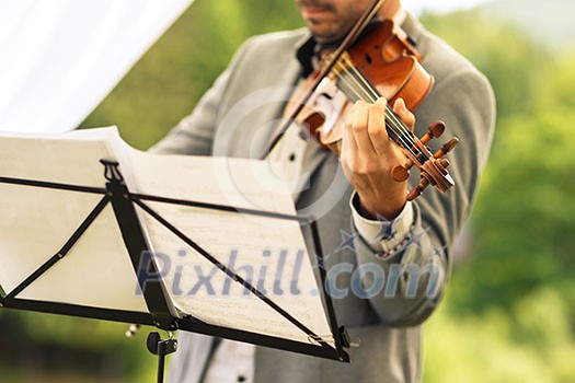Male violinist playing his instrument and reading a music sheet during an outdoor summer wedding ceremony (shallow DOF; color toned image) Male violinist playing his instrument and reading a music sheet during an outdoor summer wedding ceremony (shallow DOF; color toned image)