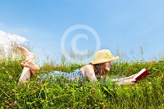 Young teenage girl reading book in summer meadow with straw hat
