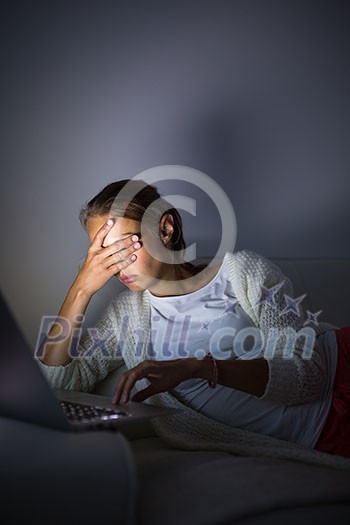 Very tired young woman, burning the midnigh oil - working late at night on her laptop computer, at home, sitting on sofa, rubbing her tired eyes, trying to stay focused