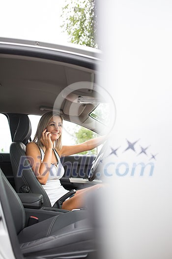 Pretty, young woman in her modern car in a parking lot, calling on her call phone, smiling, looking relaxed and happy (color toned image; shallow DOF)