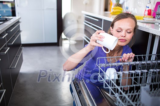 Pretty, young woman in her modern and well equiped kitchen putting cups into the dishwasher - an appliance that helps her keep the home tidy Pretty, young woman in her modern and well equiped kitchen putting cups into the dishwasher - an appliance that helps her keep the home tidy