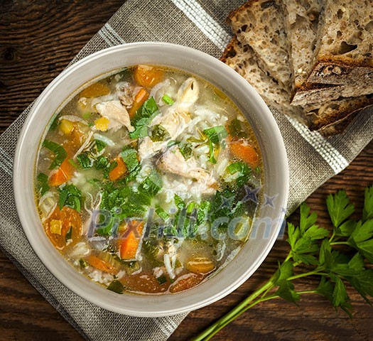 Chicken rice soup with vegetables in bowl and bread from above closeup