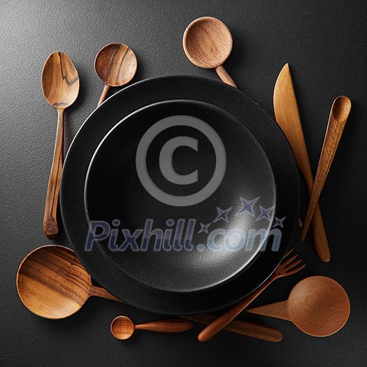 two black plates and wooden spoon, fork, knife on a black table.